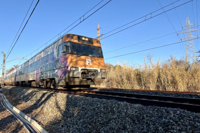 Cabecera de un tren de la red de Rodalies sobre una vía y rodeando de campo. Al fondo se ve un cielo azul y las catenarias de la red.