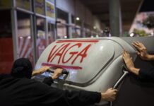 Un contenedor de basura con la palabra "vaga" (huelga) escrita en rojo y las manos de varias personas empujándolo.
