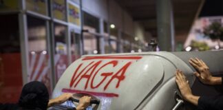 Un contenedor de basura con la palabra "vaga" (huelga) escrita en rojo y las manos de varias personas empujándolo.