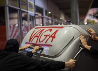 Un contenedor de basura con la palabra "vaga" (huelga) escrita en rojo y las manos de varias personas empujándolo.