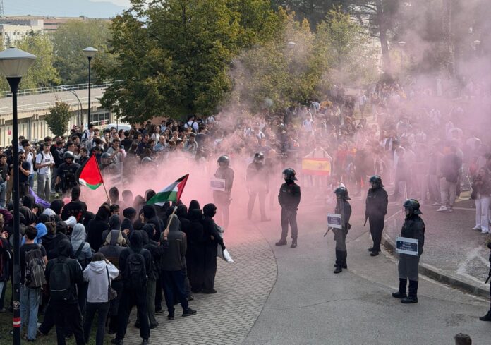 Enfrentamiento entre manifestantes con banderas palestinas y agentes antidisturbios durante una protesta con humo rosa en el ambiente.