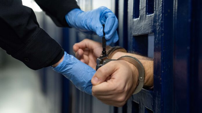 Un policía colocando las esposas a un detenido que se encuentra detrás de una reja.