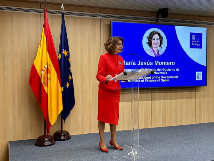 La ministra de Hacienda, María Jesús Montero, en una rueda de prensa en Bruselas, delante de las banderas de España y la UE y de una pantalla con sus datos.