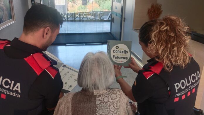 Dos agentes de los Mossos d'Esquadra muestran un folleto con consejos de seguridad a una mujer de avanzada edad para prevenir estafas.