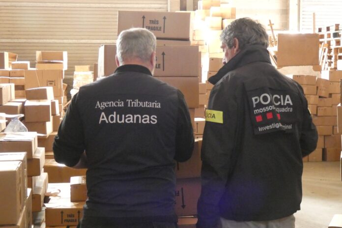 Un agente de los Mossos d’Esquadra y otro de Vigilancia Aduanera observan de espaldas miles de cajas de perfumes falsificados en una nave de La Selva.