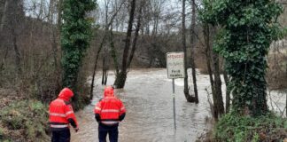 Dos efectivos de los Bombers con chaquetas reflectantes naranjas observan una carretera inundada por la crecida de un río, junto a una señal de peligro por riadas.