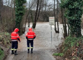 Dos efectivos de los Bombers con chaquetas reflectantes naranjas observan una carretera inundada por la crecida de un río, junto a una señal de peligro por riadas.