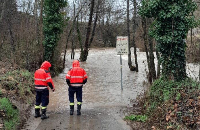 Dos efectivos de los Bombers con chaquetas reflectantes naranjas observan una carretera inundada por la crecida de un río, junto a una señal de peligro por riadas.