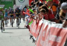 Ciclistas de competición circulando a gran velocidad por una calle urbana rodeada de vallas y público animando.