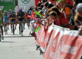 Ciclistas de competición circulando a gran velocidad por una calle urbana rodeada de vallas y público animando.