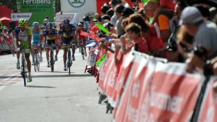 Ciclistas de competición circulando a gran velocidad por una calle urbana rodeada de vallas y público animando.