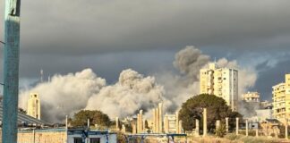 Densa columna de humo gris tras un bombardeo en una zona urbana costera de Tiro, Líbano, con civiles observando desde una plataforma en primer plano.