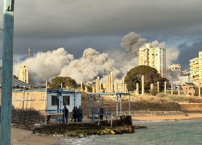 Densa columna de humo gris tras un bombardeo en una zona urbana costera de Tiro, Líbano, con civiles observando desde una plataforma en primer plano.