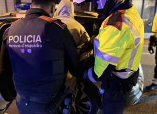 Primer plano de dos agentes de los Mossos d’Esquadra, uno de paisano con chaleco y otro con chaqueta reflectante, deteniendo a un hombre con sudadera con capucha de noche.