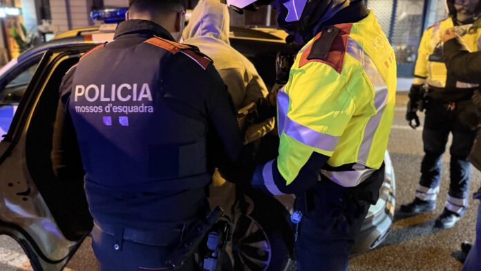 Primer plano de dos agentes de los Mossos d’Esquadra, uno de paisano con chaleco y otro con chaqueta reflectante, deteniendo a un hombre con sudadera con capucha de noche.
