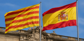 Banderas de Cataluña y España ondeando juntas frente a un edificio institucional.