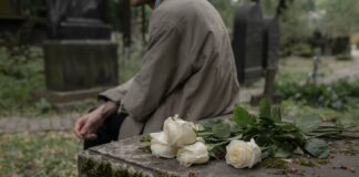 Rosas blancas sobre una tumba en un cementerio mientras una persona permanece sentada junto a la lápida.