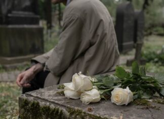 Rosas blancas sobre una tumba en un cementerio mientras una persona permanece sentada junto a la lápida.