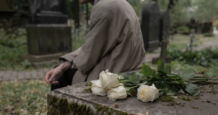 Rosas blancas sobre una tumba en un cementerio mientras una persona permanece sentada junto a la lápida.